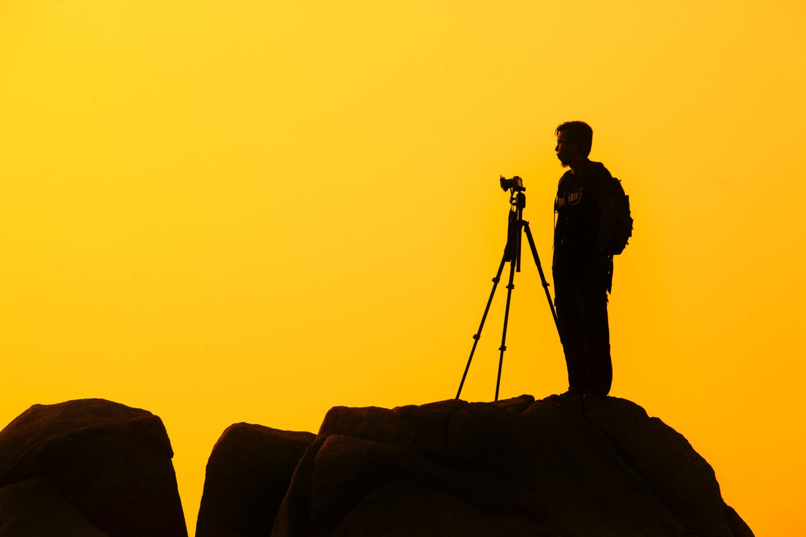 Black silhouette of a man and a camera on a tripod ontop of a cliff with a yellow background